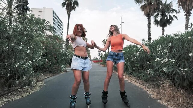 Two cheerful young women holding hands and inline skating