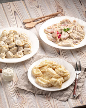 Three white plates with assorted cooked vareniki with different fillings and meat dumplings on rustic wooden background