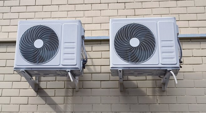 Two white outdoor air conditioning units mounted on beige brick wall with visible fans and metal brackets