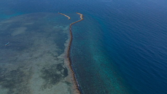 Aerial view of a long stone breakwater separating shallow turquoise waters from the deep blue sea in Indonesia.