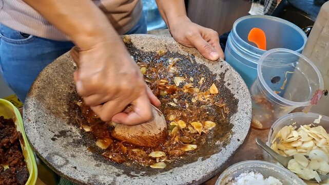 Indonesian street food vendor preparing Tahu Gejrot, a traditional West Java dish. Hands crushing garlic, shallots, and chili in an earthenware mortar to make the sweet, spicy sauce..