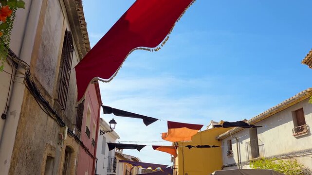 Red, orange and black festive banners fluttering in the wind above a narrow street in an old Spanish town decorated for a grape harvest festival under a bright blue sky.