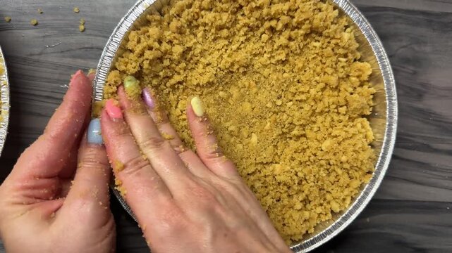 In a cozy kitchen, a woman presses fresh biscuit crumbs into a cheesecake pan with her own hands, creating a very firm and delicious base for the crust, a warm candid authentic life moment.