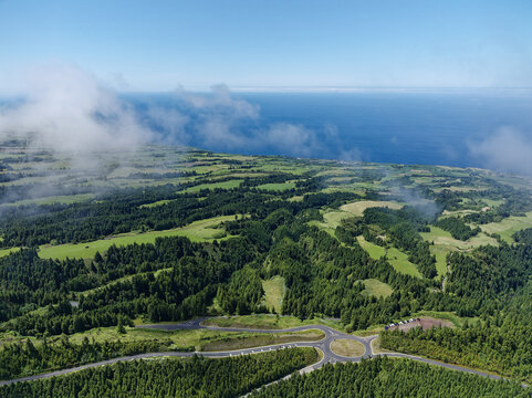 Aerial view of the lush green landscape featuring dense forests, rolling meadows, and a winding road with a roundabout near the coastline in Azores, Portugal.