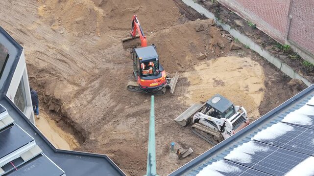 High-angle drone view of a small excavator digging brown soil between two buildings at a construction site, while a worker walks alongside during active urban construction.