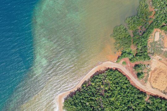 Aerial view of the PT Ceria Nugraha Indotama mining site area featuring a dirt road along the coastline, lush green vegetation, and clear turquoise waters in Wolo, South East Sulawesi, Indonesia.