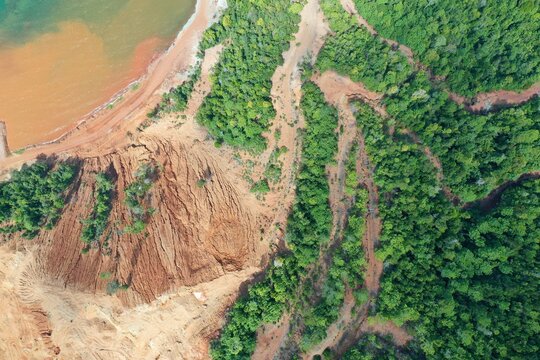 Aerial view of the PT Ceria Nugraha Indotama mining site showing exposed red earth, excavation areas, and lush tropical forest meeting the coastline in Wolo, South East Sulawesi, Indonesia.