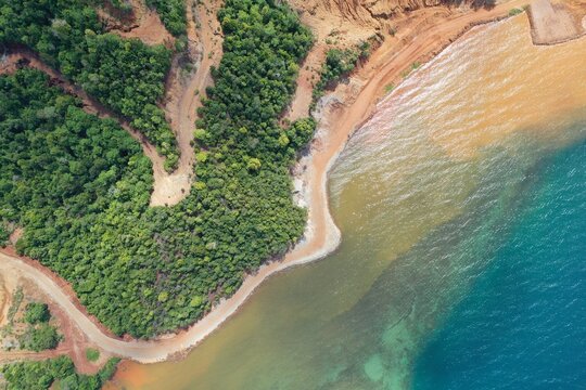 Aerial view of the PT Ceria Nugraha Indotama mining site showing lush green forest, dirt access roads, and sediment-rich water along the coastline in Wolo, South East Sulawesi, Indonesia.