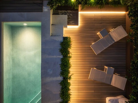 Aerial view of a modern swimming pool and wooden deck at night, featuring illuminated lounge chairs, lush greenery, and warm ambient lighting in San Benedetto del Tronto, Marche, Italy.
