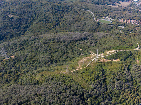 Aerial view of power lines traversing a lush green forested landscape with a dirt road and a small sports field in the distance in Sariyer, Istanbul, Turkey.
