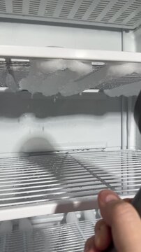 Home freezer defrosting: owner aims hair dryer at thick ice, chunks melt and drop, water collects, candid authentic life chore captured in real kitchen with towels and bucket today.