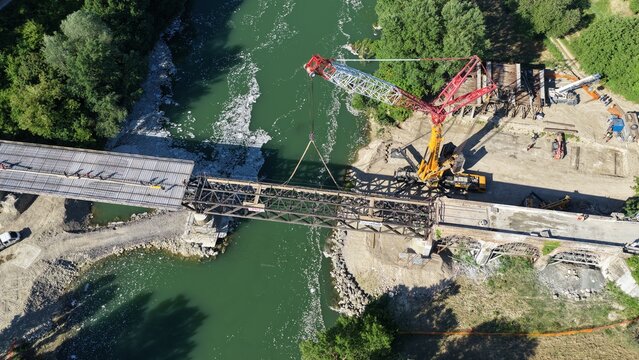 Aerial view of a bridge construction site with a large crane lifting a steel truss section over a river in Todi, Umbria, Italy.