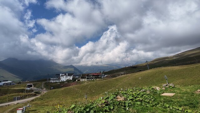 Scenic view of a mountain village with white houses scattered on a green hillside, surrounded by rolling hills and dramatic clouds over distant mountain peaks.