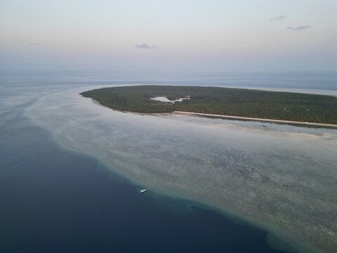 Aerial view of Moromaho Island surrounded by clear turquoise waters, coral reefs, and a small boat in the deep blue sea near Binongko, South East Sulawesi, Indonesia.