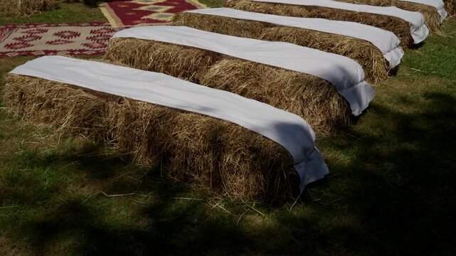 Hay bale seating rows covered with white fabric on a lawn with patterned rugs for a rustic ceremony