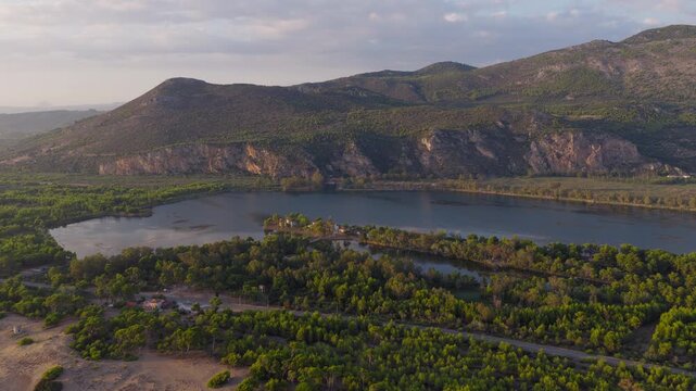 Aerial footage of Kaiafas lake near Zacharo, Ilia, Greece at sunset, with serene water, lush green forest, distant rugged mountains, golden hour lighting.