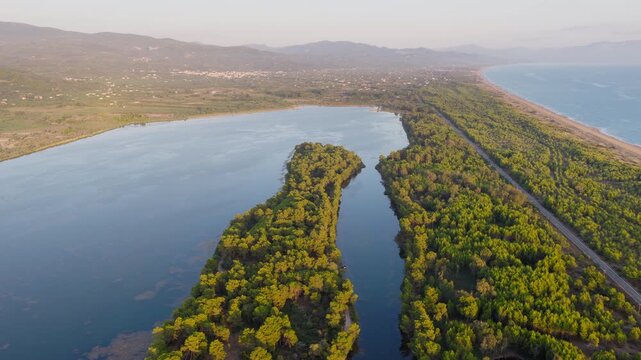 Stunning drone video captures Kaiafas Lake and its lush wetlands in Zacharo, Ilia, Greece at golden hour. Sunlight glows over tranquil water, green forest, and coastal landscape. No people visible.