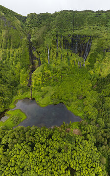 Aerial view of the lush green cliffs and multiple waterfalls cascading into a dark lake at Poco da Alagoinha on Flores Island in Faja Grande, Azores, Portugal.
