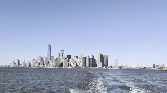View from the stern of the Staten Island Ferry moving on dark blue water, leaving a white foam wake. Blue sky with seagulls, and the Downtown Manhattan skyline and Brooklyn Bridge in the background.