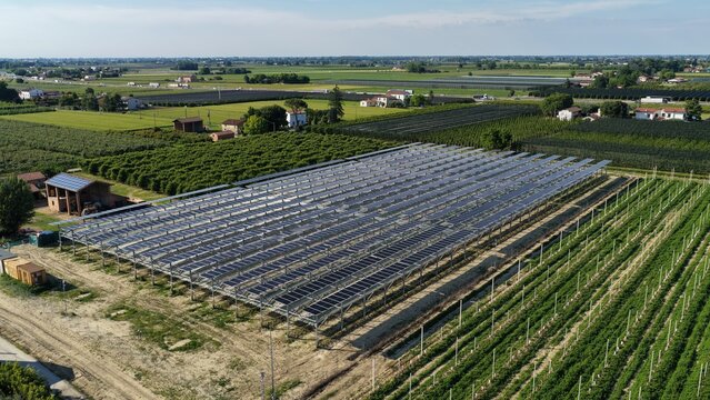 Aerial view of a large solar farm with rows of photovoltaic panels installed in a rural agricultural landscape surrounded by green orchards and fields in Faenza, Emilia-Romagna, Italy.