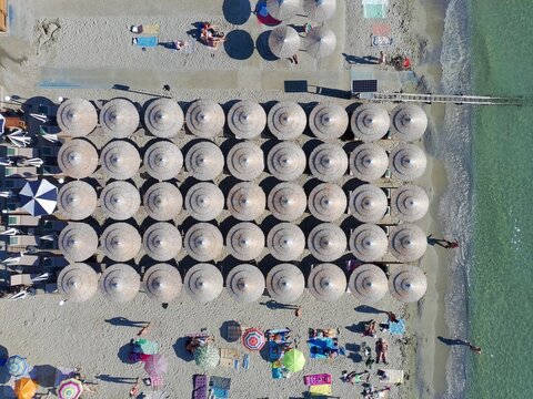 Aerial view of rows of straw umbrellas and sun loungers arranged on the sandy beach along the turquoise water in Katarinies Paralia, Greece.