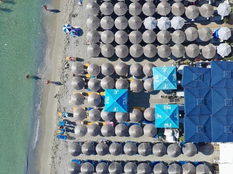 Aerial view of the beach with rows of straw umbrellas, blue square umbrellas, and clear turquoise water along the sandy coastline in Katarinies Paralia, Greece.