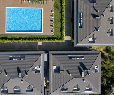 Aerial view of a swimming pool with blue water surrounded by a wooden deck and green hedges, adjacent to modern residential buildings with grey tiled roofs in Bursa, Bursa, Turkiye.