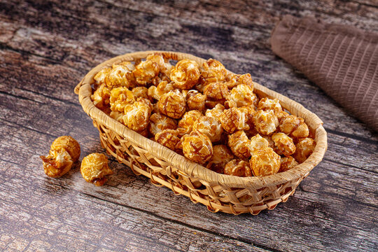 Caramel popcorn in woven basket on rustic wooden surface with scattered kernels and brown cloth background