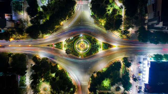 Aerial view of an illuminated roundabout in Surabaya with light trails from moving vehicles, surrounded by lush trees and urban infrastructure in Surabaya, East Java, Indonesia.