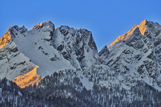 Aerial view of Valchiavenna mountains covered in snow with golden hour sunlight hitting the peaks and pine forests below in Lombardy, Italy.