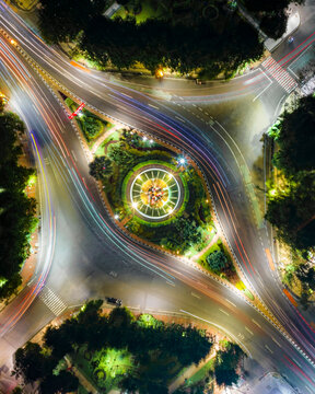 Aerial view of the illuminated roundabout with vibrant light trails from night traffic, lush surrounding greenery, and urban road infrastructure in Surabaya, East Java, Indonesia.
