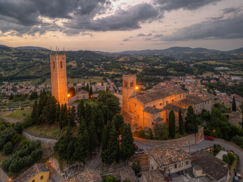 Aerial view of the historic Duomo Vecchio and Torre degli Smeducci illuminated at dusk, surrounded by cypress trees and overlooking the town of San Severino Marche, Marche, Italy.