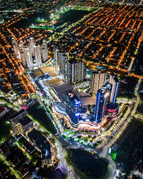 Aerial view of the illuminated cityscape at night, featuring modern high-rise apartment complexes, a large shopping mall, and glowing street lights in Surabaya, East Java, Indonesia.