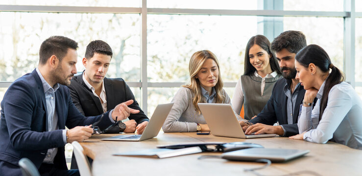 Business executives sitting around conference table in office explaining corporate project details on laptops.