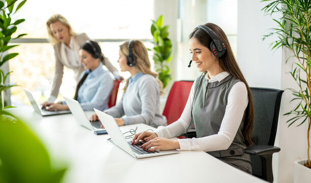 Customer support team with headsets typing on laptops while female manager assists in office.