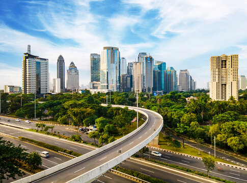 Aerial view of the modern Jakarta skyline featuring towering skyscrapers, lush green trees, and a curved elevated highway under a bright blue sky in Jakarta, Jakarta, Indonesia.