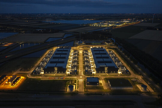 Aerial view of a large illuminated hyperscale data center complex at night, featuring rows of server buildings, internal roads, and surrounding dark agricultural fields.