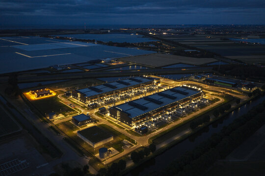 Aerial view of a modern hyperscale data center illuminated at night, featuring large server buildings, cooling infrastructure, and surrounding rural landscape.