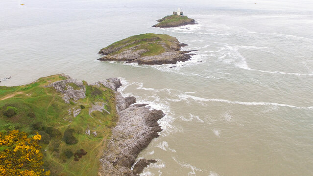 Aerial view of Mumbles Lighthouse on a rocky island surrounded by sea with rugged coastal cliffs and green hills in the foreground Mumbles, Wales, United Kingdom.
