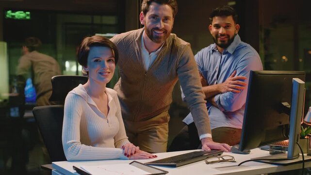 Three multiethnic business colleagues talking smiling looking at camera at dark night office diversity coworkers team talk discussing computer deadline work smile joyful posing at late evening company