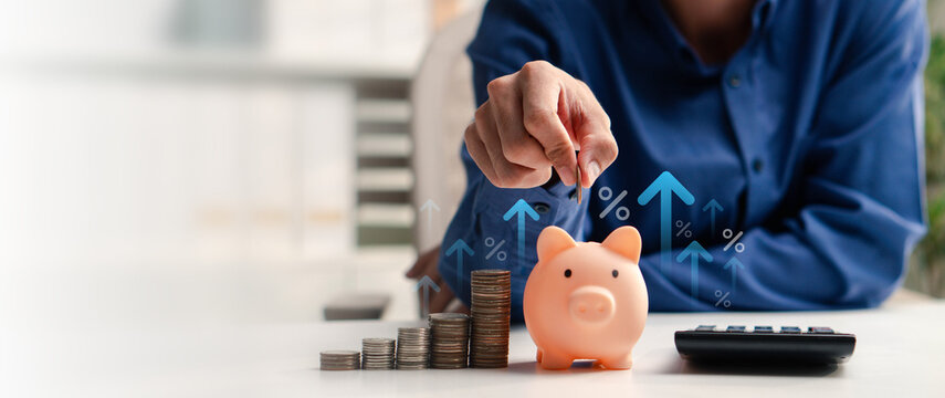 Person inserts a coin into a piggy bank beside rising coin stacks. Growth arrows and percent symbols illustrate savings, investment returns, interest rates, budgeting, and financial planning.