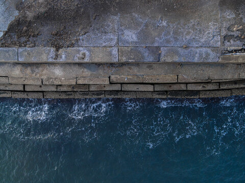 Aerial view of a concrete seawall meeting the deep blue ocean water, showing textured stone blocks and gentle waves along the coast, Raja Ampat, Southwest Papua, Indonesia.