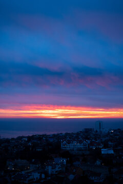 vibrant sunrise over sea and coastal town vertical shot, dramatic orange and purple sunset clouds over ocean and city skyline, panoramic view of sea horizon with glowing twilight sky