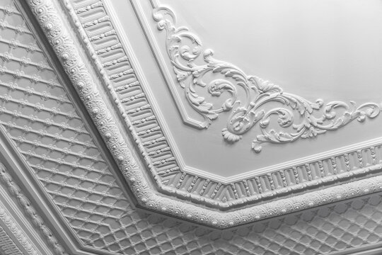 Elegant white plaster ceiling featuring ornate scrollwork, decorative molding and repeating geometric panels; conveys historic charm, craftsmanship and refined architectural details