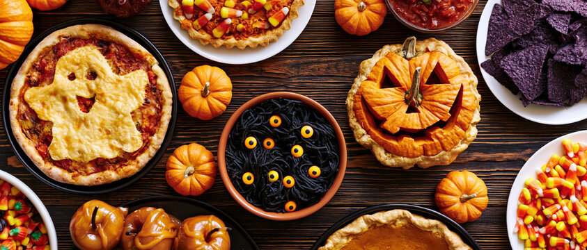 A festive spread of Halloween-themed foods, including a ghost-shaped pizza, jack-o'-lantern stuffed pumpkin, and pumpkin pie, arranged on a rustic wooden table.