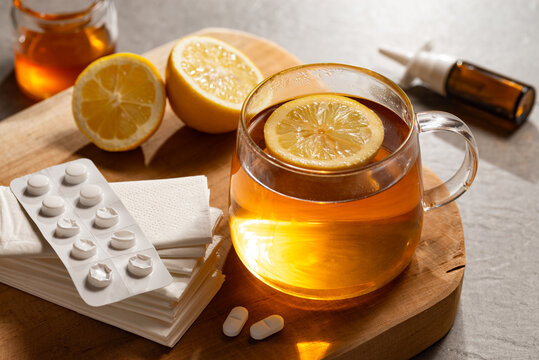 Erk&auml;ltungszeit - Hei&szlig;er Tee in Glastasse auf Holzbrett mit Zitronen, Nasenspray und Tabletten