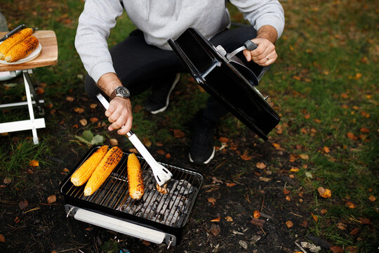 Cropped closeup of man cooking sweet corn cobs on portable charcoal grill with tongs at campsite in autumn forest, holding barbecue lid. Outdoor kitchen, camp food, picnic picnic, autumn vibe