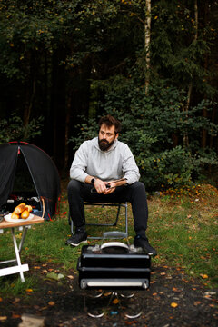 Bearded man sitting on chair at campsite in autumn forest, waiting for food to cook on portable barbecue grill near tent and table. Camp life, outdoor relaxation, hiking, eco tourism, lifestyle, verti
