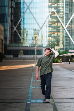 A young man walks along an open pedestrian path, adjusting his hair. He is wearing a loose olive-green T-shirt and dark trousers. The scene takes place in a city.