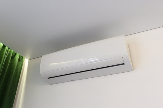 Sleek white wall-mounted split-system air conditioner under ceiling, smooth glossy finish, horizontal air outlet, clean minimalist design, adjacent green velvet curtain